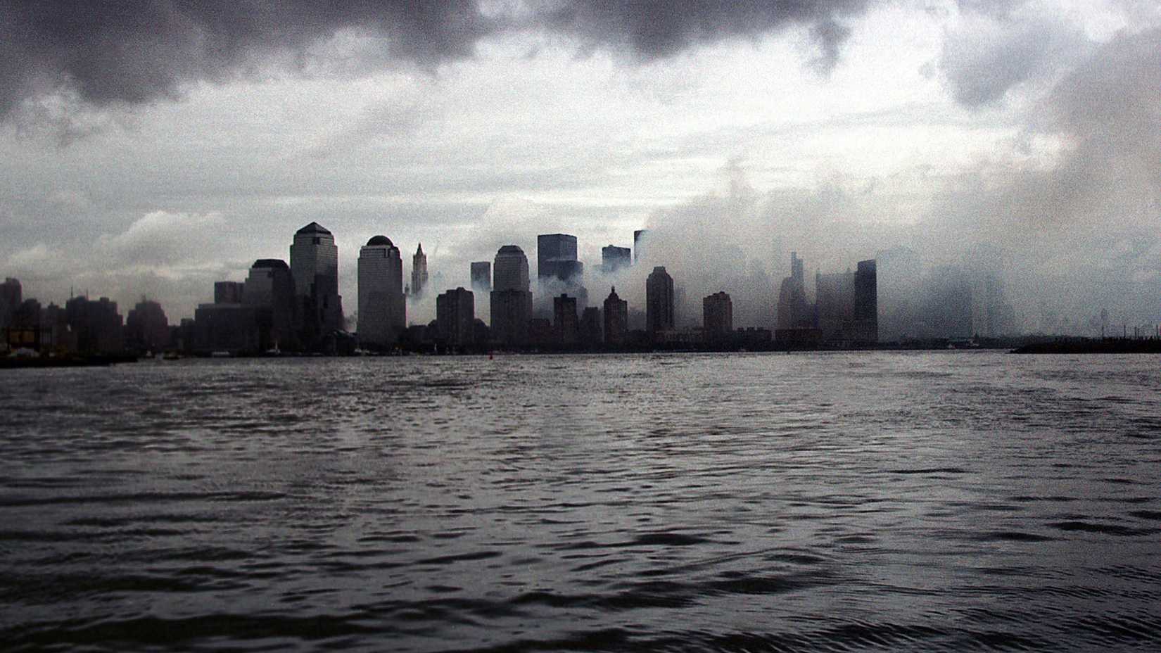 Smoke hangs over Lower Manhattan beneath a cloudy sky, viewed across the Hudson River from Liberty State Park.