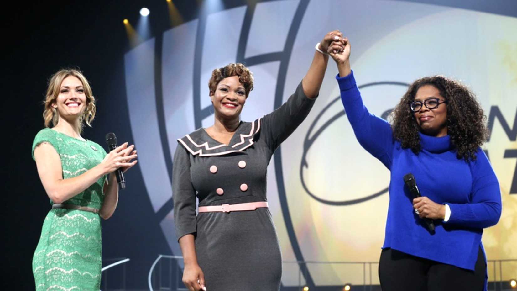 Jas Boothe, center, and Oprah Winfrey raise their clasped hands onstage while a woman in a green dress applauds.