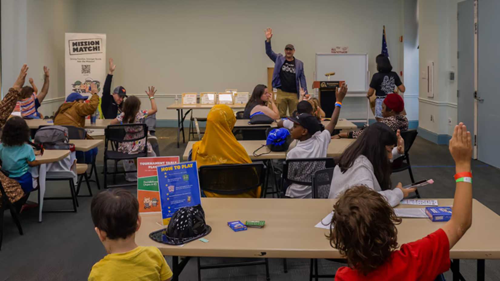 Children seated at tables with Mission Match cards raise their hands as a presenter raises his hand at the front.